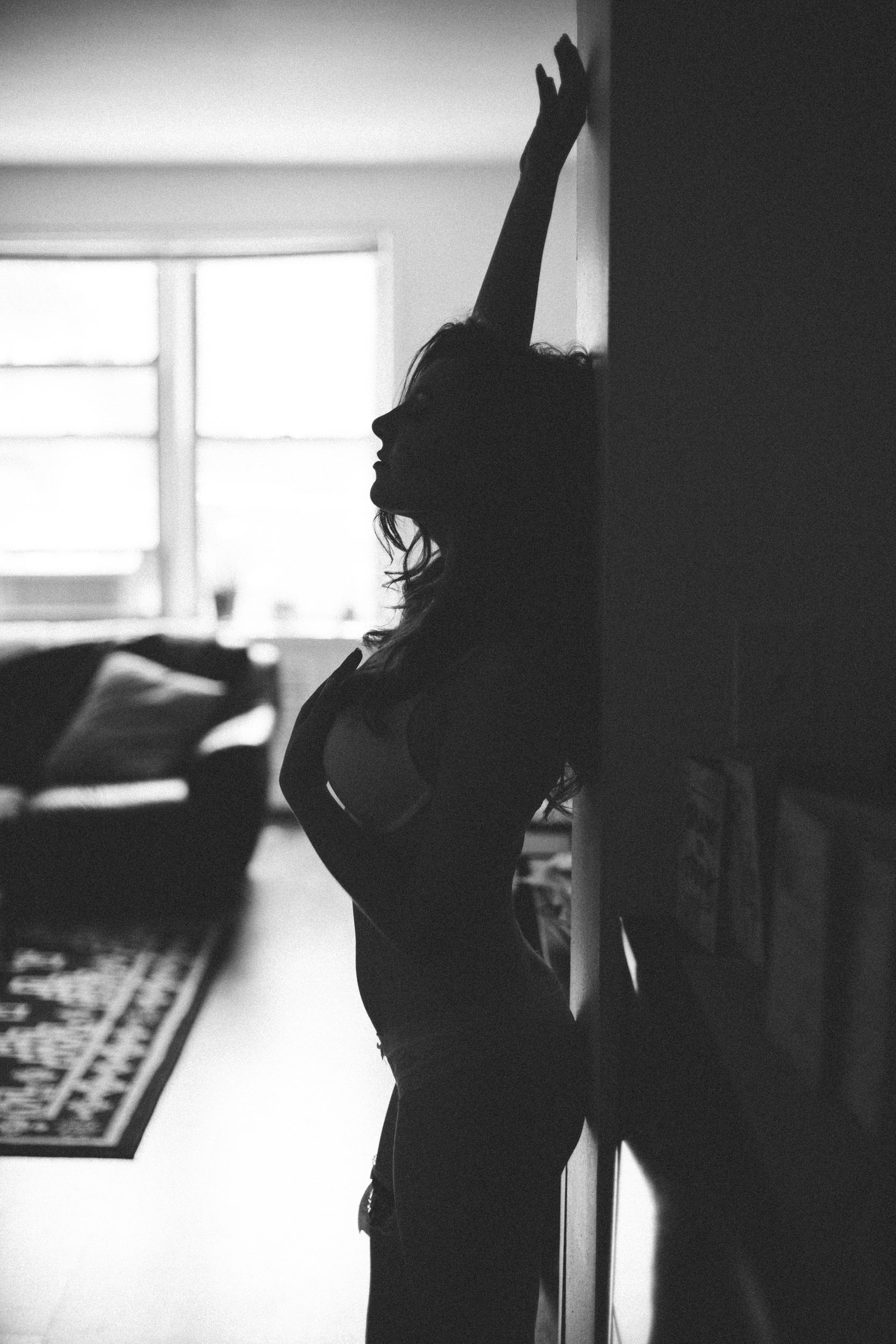 Black and white silhoutte portrait of a woman during her boudoir photoshoot. Leaning against a wall with her hand above her head.